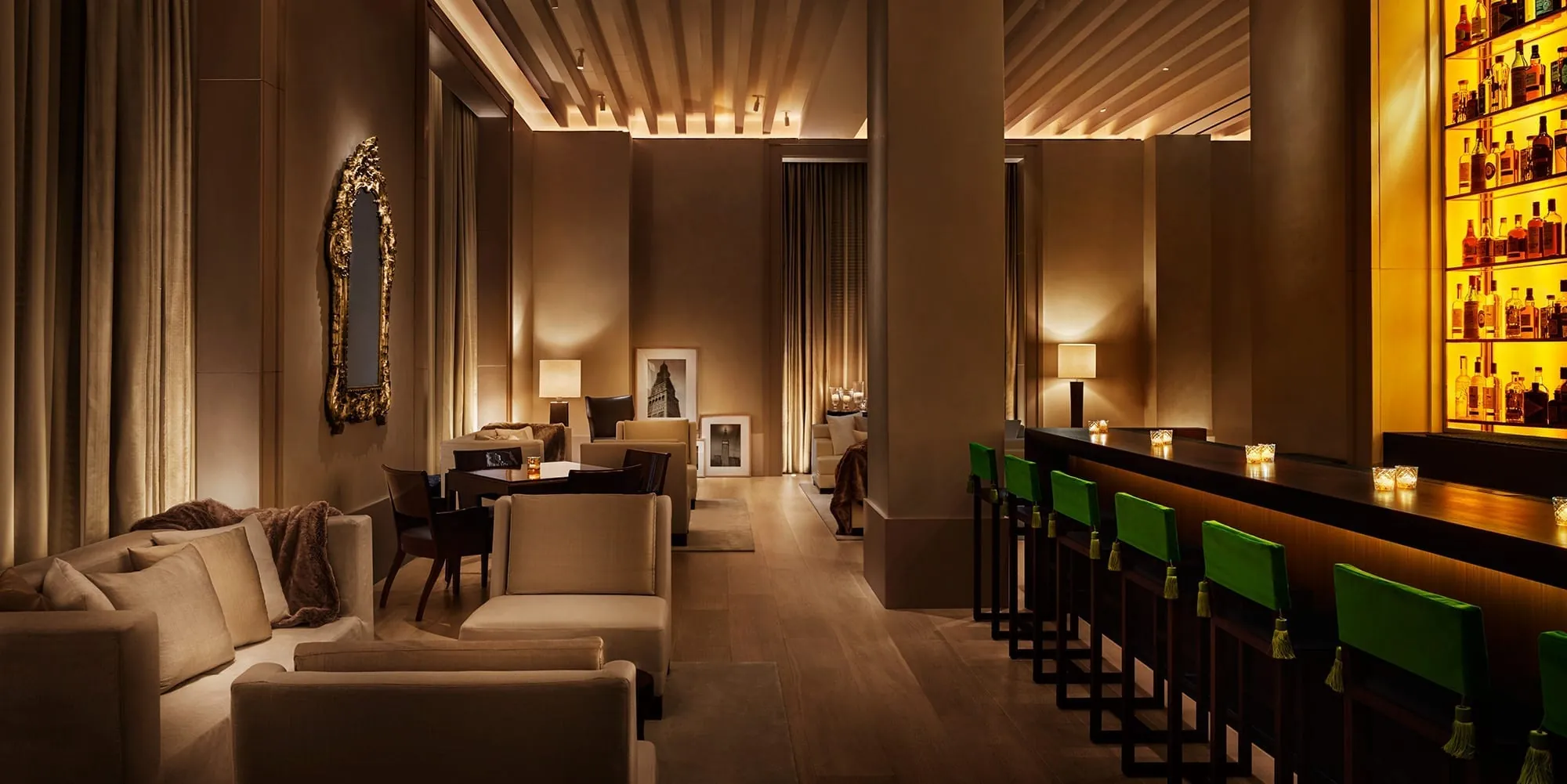 A candlelit hotel lounge bar, cream sofas along one wall and green velvet stools before a glowing back bar.