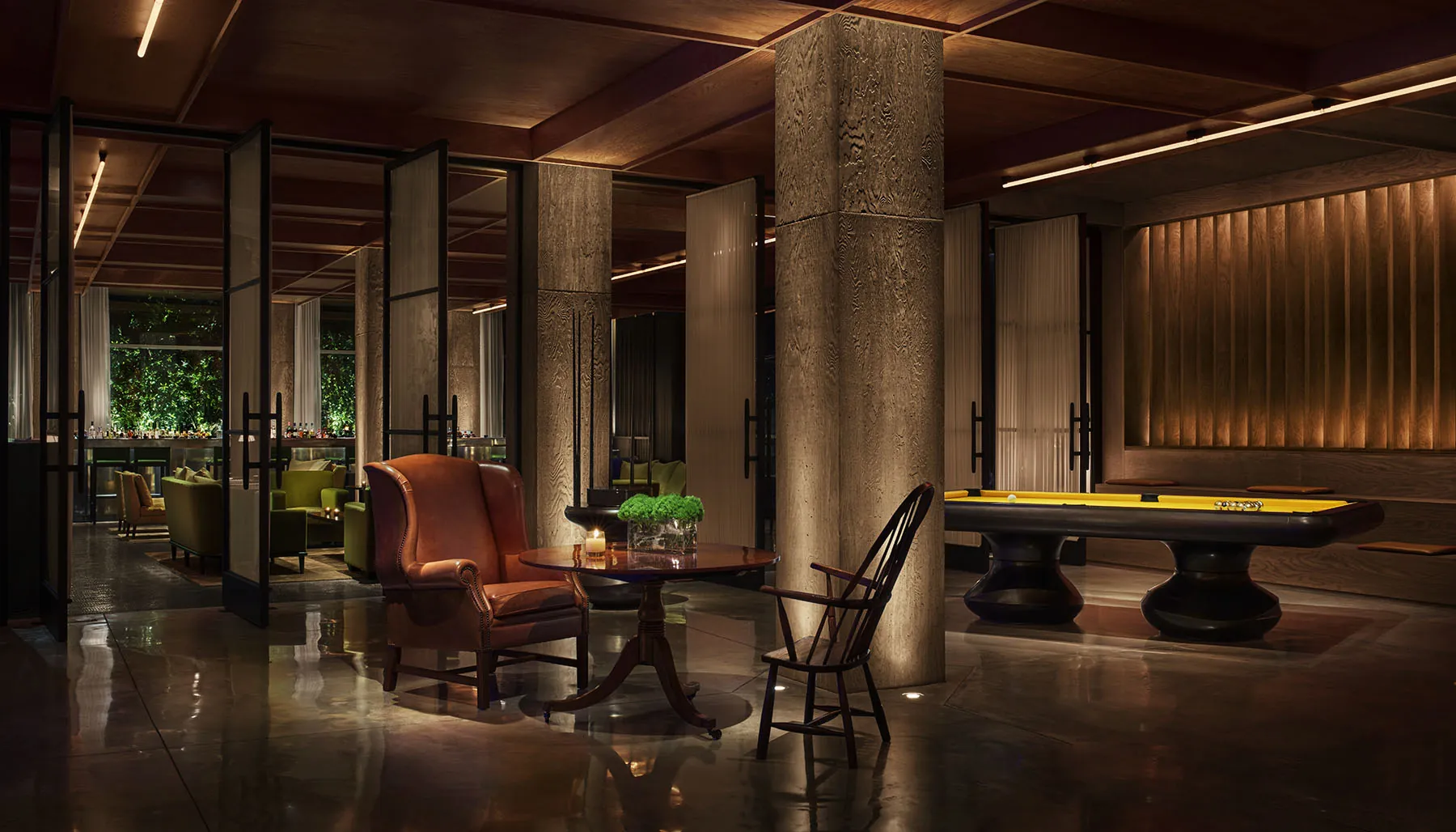 A low-lit hotel lounge with a leather wingback chair beside a candlelit table, concrete columns, and a pool table in warm light.